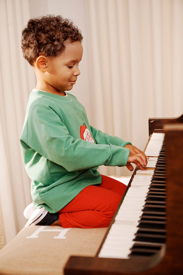 A young child in a Kiawah Kelly Green Cassidy Comfy Crewneck with Santa by The Beaufort Bonnet Company and red pants sits on a piano bench, playing the keys with both hands. The child is focused on the piano, while light curtains in the background create a cozy scene perfect for Christmas gatherings.