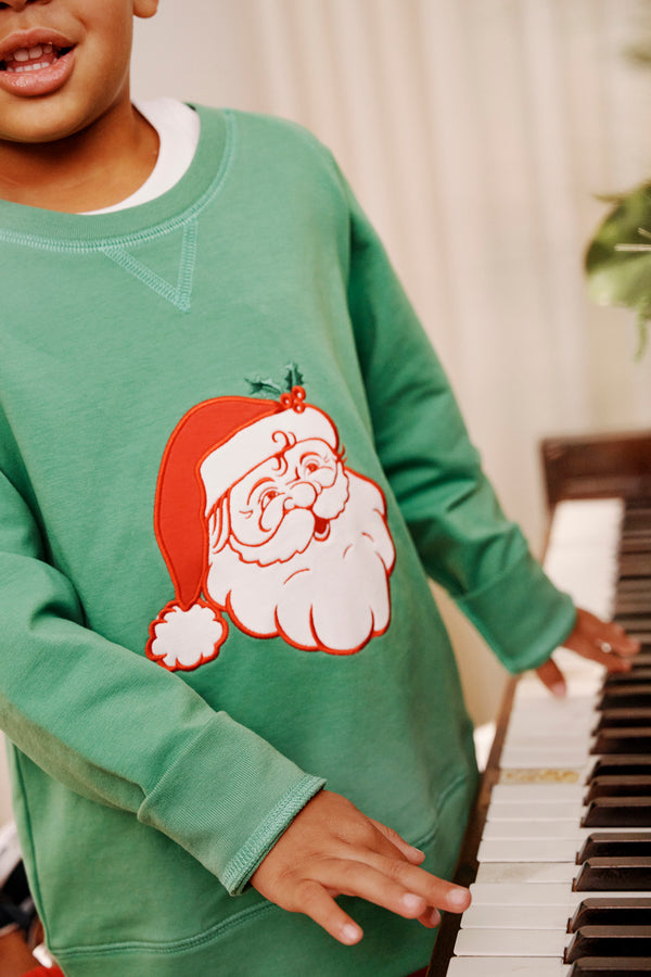 A child wearing The Beaufort Bonnet Company’s Cassidy Comfy Crewneck in Kiawah Kelly Green with a Santa motif stands by a piano, placing their hands on the keys.