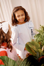 Two young girls stand together indoors, smiling and chatting. One girl is wearing The Beaufort Bonnet Company’s Dani Dress in Buckhead Blue Corduroy, while the other wears a red and pink outfit with a white hair bow. Green plants are visible in the foreground.