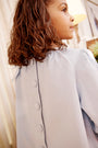 A young girl with curly brown hair is standing indoors, looking to the side, wearing The Beaufort Bonnet Company’s Dani Dress in Buckhead Blue Corduroy with buttons down the back.