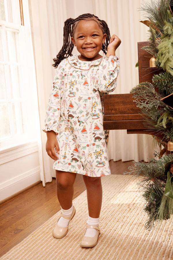 A young girl wearing The Beaufort Bonnet Company’s Long Sleeve Ramona Ruffle Dress - 12 Days of Christmas and beige shoes smiles as she stands by a piano adorned with holiday greenery in a bright room with wooden floors and white curtains.