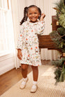 A young girl wearing The Beaufort Bonnet Company’s Long Sleeve Ramona Ruffle Dress - 12 Days of Christmas and beige shoes smiles as she stands by a piano adorned with holiday greenery in a bright room with wooden floors and white curtains.