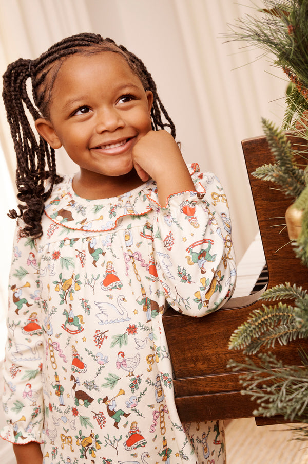 A young girl with braided hair smiles while standing next to a piano and a decorated Christmas tree, wearing The Beaufort Bonnet Company’s Long Sleeve Ramona Ruffle Dress - 12 Days of Christmas.