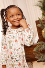 A young girl with braided hair smiles while standing next to a piano and a decorated Christmas tree, wearing The Beaufort Bonnet Company’s Long Sleeve Ramona Ruffle Dress - 12 Days of Christmas.