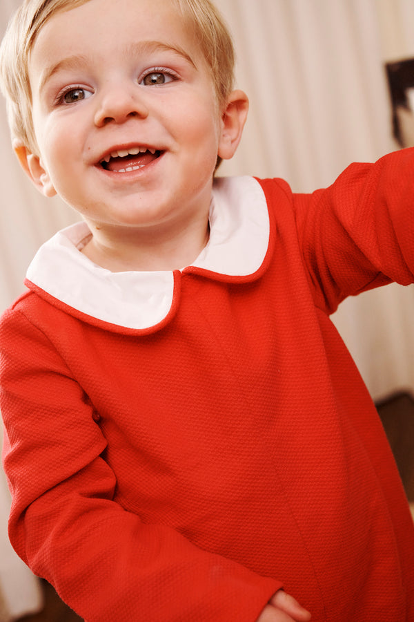 A smiling young child with light hair wears The Beaufort Bonnet Company’s John Hall Jon Jon in Richmond Red Pique & Worth Avenue White, featuring a classic white Peter Pan collar, while standing indoors against a light-colored background.