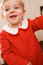 A smiling young child with light hair wears The Beaufort Bonnet Company’s John Hall Jon Jon in Richmond Red Pique & Worth Avenue White, featuring a classic white Peter Pan collar, while standing indoors against a light-colored background.
