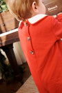 A young child wearing The Beaufort Bonnet Company’s John Hall Jon Jon in Richmond Red Pique & Worth Avenue White, featuring a white collar and back buttons, stands in front of a wooden piano with the name Howard visible on it.