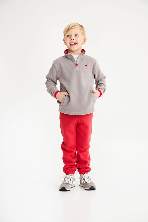 A smiling young boy wears a Hayword Half-Zip (Fleece) - Grantley Gray with Richmond Red, paired with red sweatpants and gray sneakers.