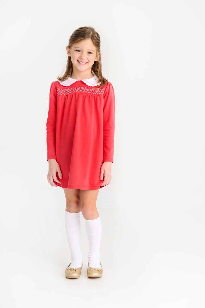 A young girl smiles while wearing a Long Sleeve Smocked Holly Day Dress - Richmond Red with Worth Avenue White, styled with a white collared neckline, white knee-high socks, and gold flats.