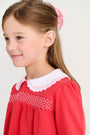 A young girl wears a Long Sleeve Smocked Holly Day Dress - Richmond Red with Worth Avenue White, featuring a white scalloped collar and smocked detailing across the chest.