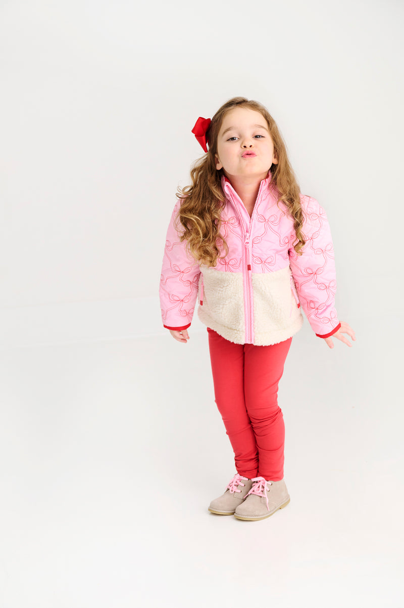 A young girl with long, curly hair, wearing a pink jacket, a red bow, and The Beaufort Bonnet Company’s Mitzy Sue Slacks in Richmond Red, stands against a white background making a playful pouty face.