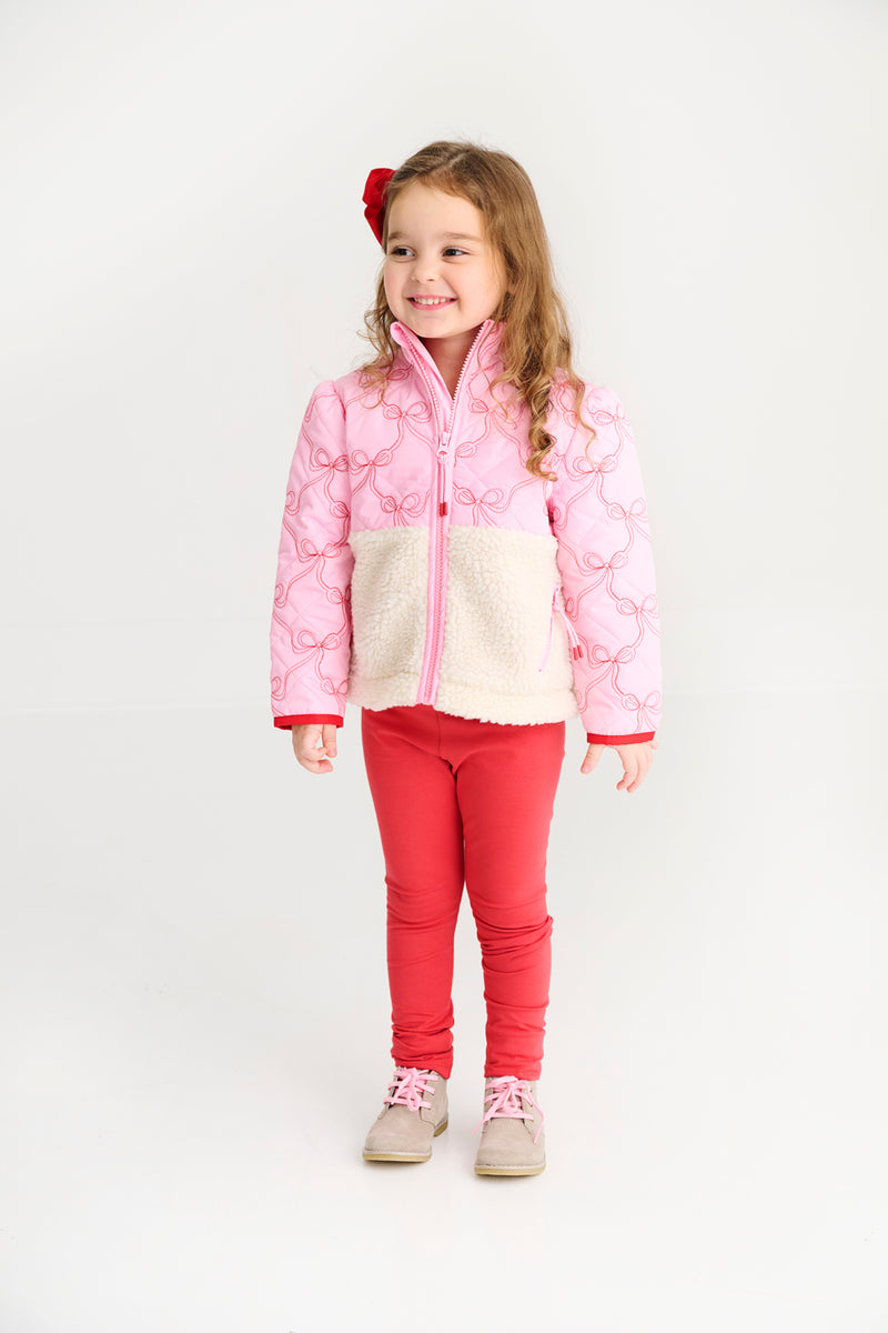 A young girl with long hair stands smiling against a plain white background, wearing The Beaufort Bonnet Company’s Cate Coat in Bluffton Bows with Palmetto Pearl, styled with red leggings, beige shoes, and a red bow with an embroidered bow pattern in her hair.