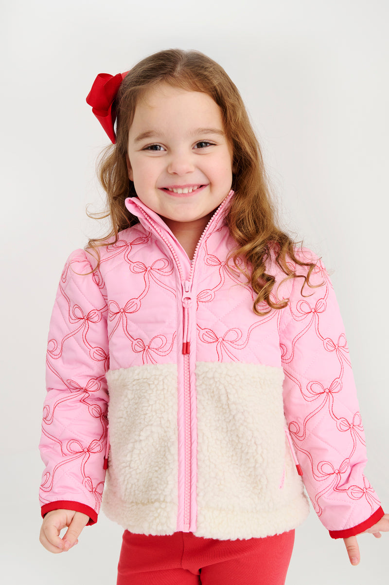 A smiling young girl with long brown hair wears The Beaufort Bonnet Company’s Cate Coat - Bluffton Bows with Palmetto Pearl, featuring a pink and white quilted design with an embroidered bow pattern; she pairs it with red pants and a matching red bow in her hair, standing against a plain white background.