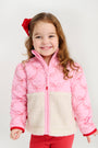 A smiling young girl with long brown hair wears The Beaufort Bonnet Company’s Cate Coat - Bluffton Bows with Palmetto Pearl, featuring a pink and white quilted design with an embroidered bow pattern; she pairs it with red pants and a matching red bow in her hair, standing against a plain white background.