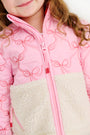 A young child is seen wearing The Beaufort Bonnet Company’s Cate Coat in Bluffton Bows with Palmetto Pearl—a quilted girls’ coat featuring an embroidered bow pattern and a cream-colored sherpa fleece panel. Only the lower half of her face and upper torso are visible as she smiles softly.