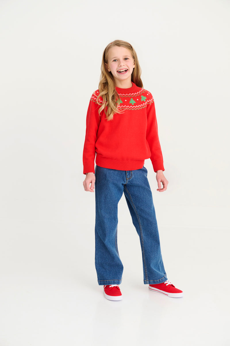 Smiling girl with long blonde hair wearing The Beaufort Bonnet Company’s Isaac’s Fair Isle Sweater in Richmond Red with Christmas Trees, paired with blue jeans and red sneakers, stands against a plain white background.