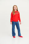 Smiling girl with long blonde hair wearing The Beaufort Bonnet Company’s Isaac’s Fair Isle Sweater in Richmond Red with Christmas Trees, paired with blue jeans and red sneakers, stands against a plain white background.
