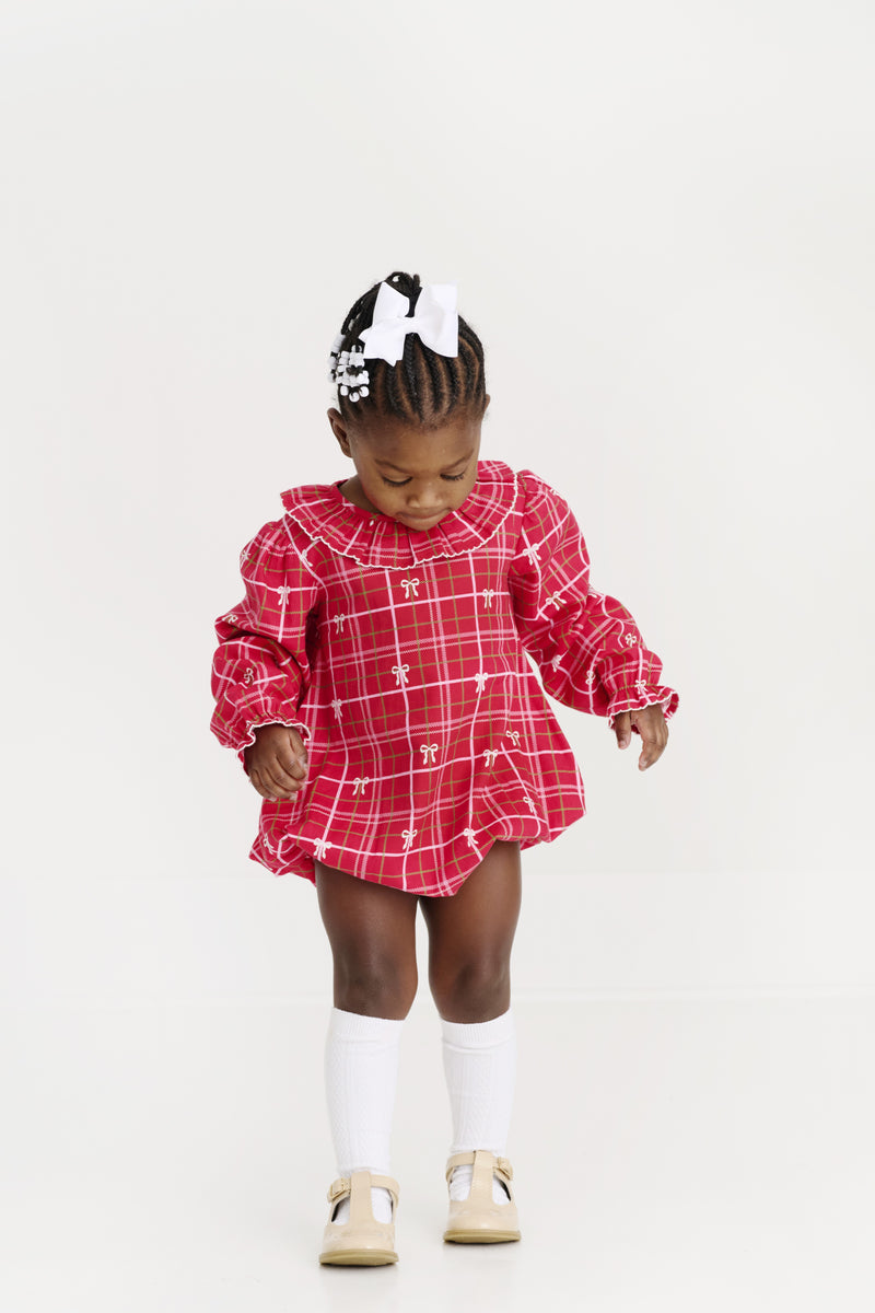 A young child wearing The Beaufort Bonnet Company’s Long Sleeve Ramona Ruffle Bubble in Torrington Tartan with Palmetto Pearl, paired with white bows, white knee-high socks, beige shoes, and a white hair bow, stands looking down against a plain white background—showcasing a charming holiday outfit.