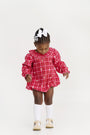 A young child wearing The Beaufort Bonnet Company’s Long Sleeve Ramona Ruffle Bubble in Torrington Tartan with Palmetto Pearl, paired with white bows, white knee-high socks, beige shoes, and a white hair bow, stands looking down against a plain white background—showcasing a charming holiday outfit.