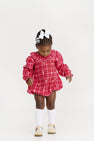 A young child wearing The Beaufort Bonnet Company’s Long Sleeve Ramona Ruffle Bubble in Torrington Tartan with Palmetto Pearl, paired with white bows, white knee-high socks, beige shoes, and a white hair bow, stands looking down against a plain white background—showcasing a charming holiday outfit.