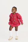 A young girl smiles brightly, wearing The Beaufort Bonnet Company’s Long Sleeve Ramona Ruffle Bubble in Torrington Tartan with Palmetto Pearl, paired with white knee-high socks, beige shoes, and a white bow in her hair, standing against a white background.