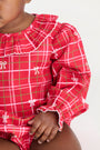 A baby wearing The Beaufort Bonnet Company’s Long Sleeve Ramona Ruffle Bubble in Torrington Tartan with Palmetto Pearl—featuring a red, white, and green plaid design with small white bows—sits against a plain light background. Only the lower face, arm, and part of the upper body are visible.
