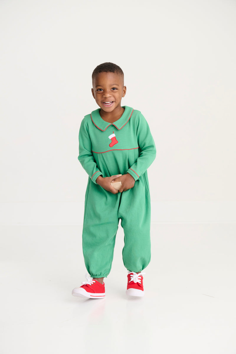 A young child smiles while standing in The Beaufort Bonnet Company’s Long Sleeve Bradford Romper - Kiawah Kelly Green with Stocking Embroidery, paired with red sneakers, against a plain white background.