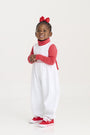 A young child smiles, wearing a red headband with a bow, a red long-sleeve top, and The Beaufort Bonnet Company’s Sleeveless Rebecca Romper in Kiawah Kelly Green Microdot with Richmond Red, crafted from soft Pima Cotton. Red sneakers complete this adorable holiday outfit as the child stands against a plain white background.