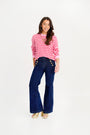A woman stands smiling on a plain white background, wearing The Beaufort Bonnet Company’s Women’s Cissy Sweater in Pier Party Pink & Richmond Red Stripe. She pairs the festive cotton sweater with dark blue high-waisted wide-leg jeans featuring gold buttons and finishes her look with gold shoes.