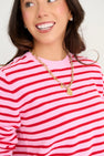 A woman smiles against a plain light background, wearing The Beaufort Bonnet Company’s Women’s Cissy Sweater in Pier Party Pink & Richmond Red Stripe along with a chunky gold charm necklace. Only her upper body and part of her face are visible.