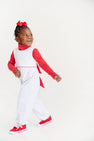A young child smiles while walking, wearing a red bow, red long-sleeve shirt, The Beaufort Bonnet Company’s Sleeveless Rebecca Romper in Kiawah Kelly Green Microdot with Richmond Red made from soft Pima Cotton, and red sneakers against a plain white background—perfect as a festive Holiday Outfit.