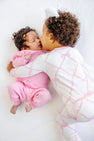 A toddler with curly hair hugs and kisses a sleeping baby, both nestled in cozy Noelle’s Night Night pajamas in the Belle Meade Bow with Pier Party Pink from The Beaufort Bonnet Company, lying on a white blanket—a true bedtime essential.
