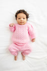 A baby with curly dark hair lies on a white blanket, wearing The Beaufort Bonnet Company’s Crawford Cable Knit Set in Pier Party Pink—a cozy sweater and matching pants—looking up with one fist slightly raised.