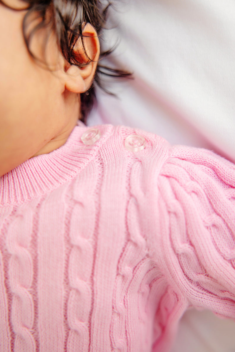 A close-up of a baby wearing The Beaufort Bonnet Company’s Crawford Cable Knit Set in Pier Party Pink, featuring clear buttons on the shoulder and showing dark curly hair and an ear.
