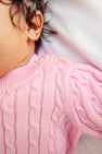 A close-up of a baby wearing The Beaufort Bonnet Company’s Crawford Cable Knit Set in Pier Party Pink, featuring clear buttons on the shoulder and showing dark curly hair and an ear.
