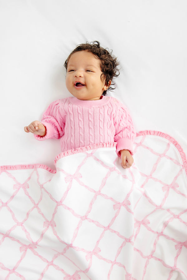A smiling baby with curly hair wearing a pink sweater lies on a white surface, partially covered by The Beaufort Bonnet Company’s Baby Buggy Blanket in the Belle Meade Bow with Pier Party Pink design—a charming white blanket featuring a pink pattern and ruffled edges—making this scene perfect for anyone searching for an ideal baby shower gift.