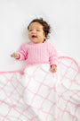 A smiling baby with curly hair wearing a pink sweater lies on a white surface, partially covered by The Beaufort Bonnet Company’s Baby Buggy Blanket in the Belle Meade Bow with Pier Party Pink design—a charming white blanket featuring a pink pattern and ruffled edges—making this scene perfect for anyone searching for an ideal baby shower gift.
