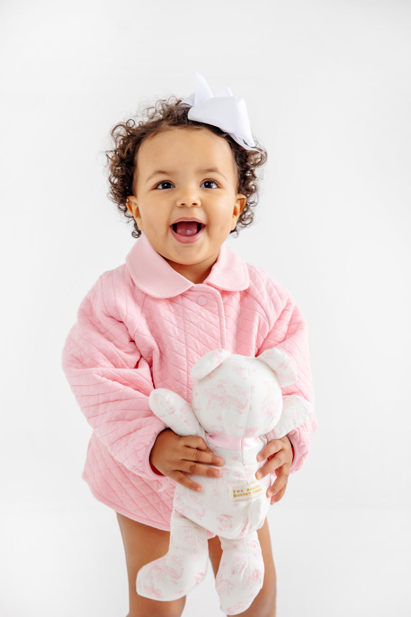 A smiling toddler with curly hair and a white bow wears a pink quilted jacket and holds The Beaufort Bonnet Company’s Bradley Bear - Sir Proper’s Sweet Arrival (Hamptons Hot Pink), a 100% cotton stuffed companion in white and pink—an adorable choice for a baby shower gift—against a plain white background.