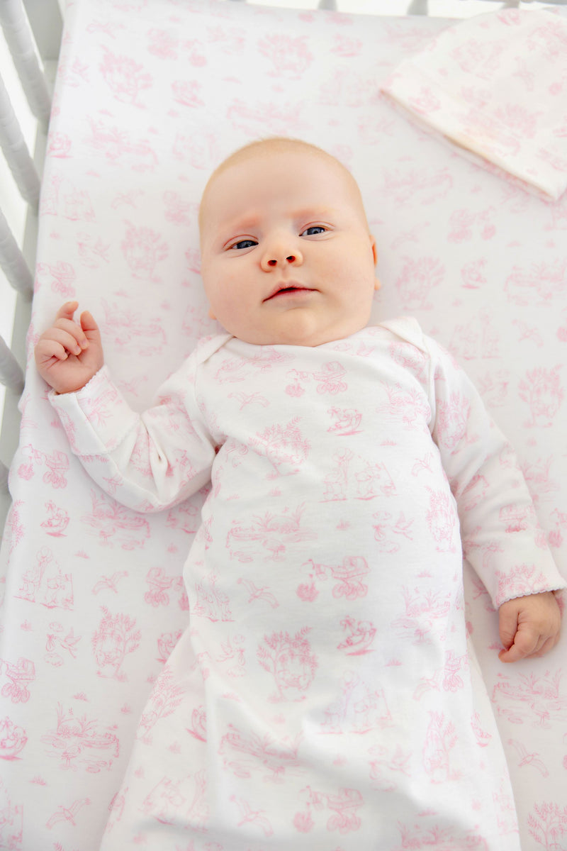 A baby with fair skin and light hair lies in a crib, wearing The Beaufort Bonnet Company’s Sir Proper’s Sweet Arrival (Hamptons Hot Pink) Bundle – Adorable Everyday Set & Bradley Bear. The soft Pima cotton layette features a charming white and pink pattern, matching the crib sheet for a coordinated look. This delightful set makes a perfect newborn bundle or thoughtful baby shower gift.
