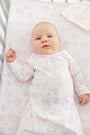 A baby with fair skin and light hair lies in a crib, wearing The Beaufort Bonnet Company’s Sir Proper’s Sweet Arrival (Hamptons Hot Pink) Bundle – Adorable Everyday Set & Bradley Bear. The soft Pima cotton layette features a charming white and pink pattern, matching the crib sheet for a coordinated look. This delightful set makes a perfect newborn bundle or thoughtful baby shower gift.
