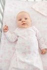 A baby with fair skin and light hair lies in a crib, wearing The Beaufort Bonnet Company’s Sir Proper’s Sweet Arrival (Hamptons Hot Pink) Bundle – Adorable Everyday Set & Bradley Bear. The soft Pima cotton layette features a charming white and pink pattern, matching the crib sheet for a coordinated look. This delightful set makes a perfect newborn bundle or thoughtful baby shower gift.