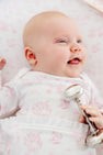 A smiling baby in The Beaufort Bonnet Company’s Sir Proper’s Sweet Arrival (Hamptons Hot Pink) Bundle—featuring an adorable everyday set and a cuddly Bradley Bear—lies on a patterned blanket, holding a silver rattle in one hand, making this the perfect newborn bundle for a memorable baby shower gift.