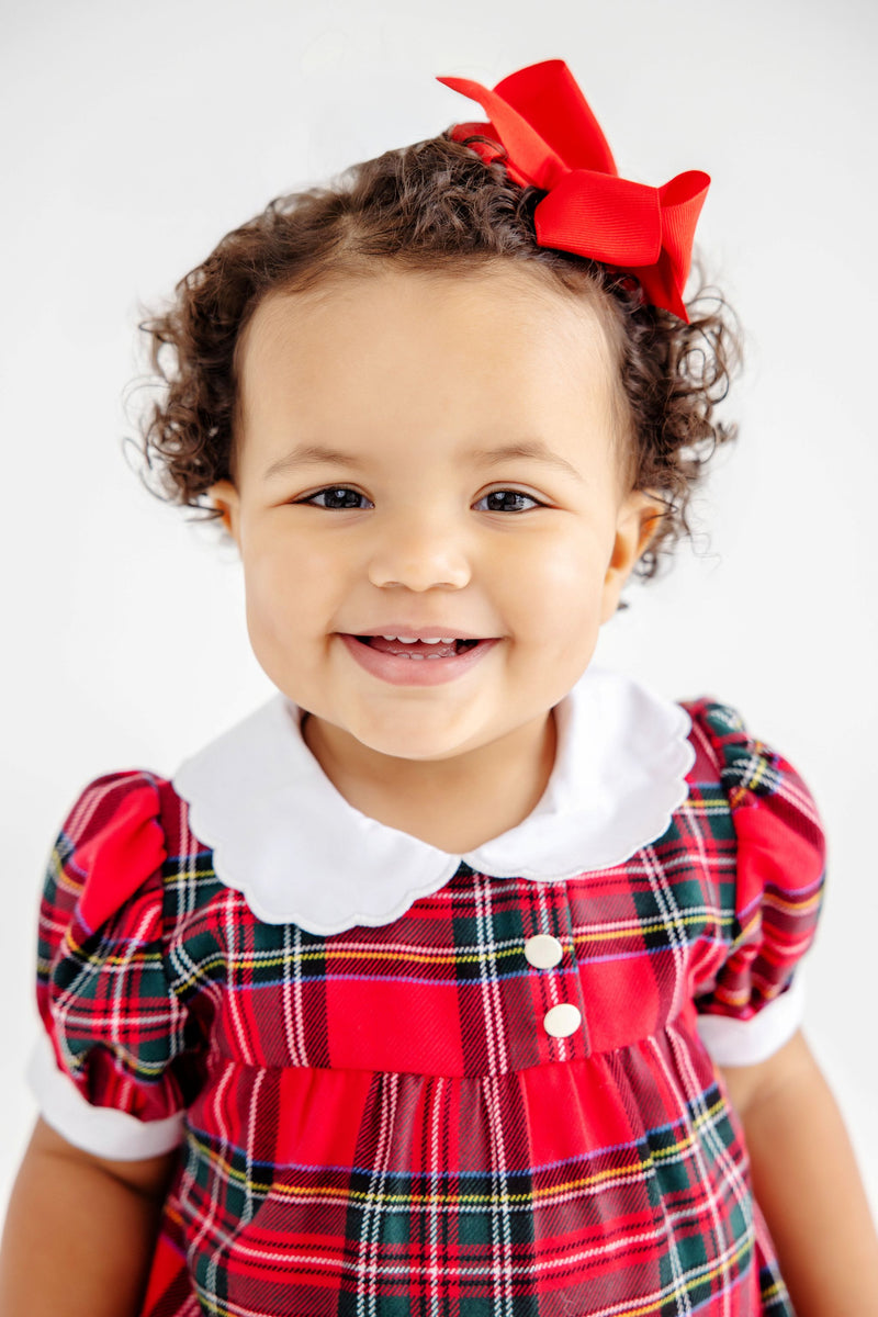 A smiling toddler wears a Mary Baker Bubble - Society Prep Plaid with Worth Avenue White, featuring a red plaid bubble romper, white scalloped collar, and matching red hair bow for a classic holiday baby outfit.