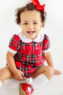 A smiling toddler wearing a Mary Baker Bubble - Society Prep Plaid with Worth Avenue White romper with a white Peter Pan collar, a red hair bow, white socks, and red shoes.