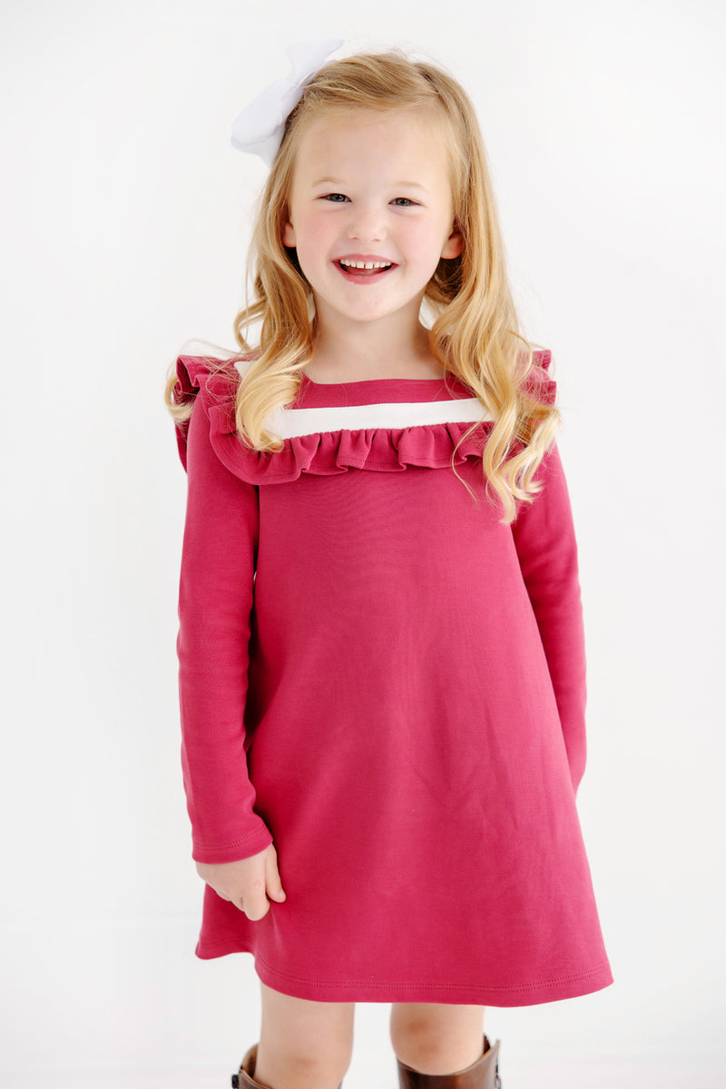 A young girl with long blonde hair and a white bow is wearing The Beaufort Bonnet Company’s Long Sleeve Darla Dress in Begley Berry with Palmetto Pearl. She is smiling and standing against a plain white background.