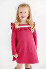 A young girl with long blonde hair and a white bow is wearing The Beaufort Bonnet Company’s Long Sleeve Darla Dress in Begley Berry with Palmetto Pearl. She is smiling and standing against a plain white background.
