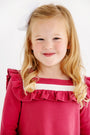 A young girl with long blonde hair, wearing a white bow and The Beaufort Bonnet Company’s Long Sleeve Darla Dress in Begley Berry with Palmetto Pearl, smiles at the camera against a plain white background.