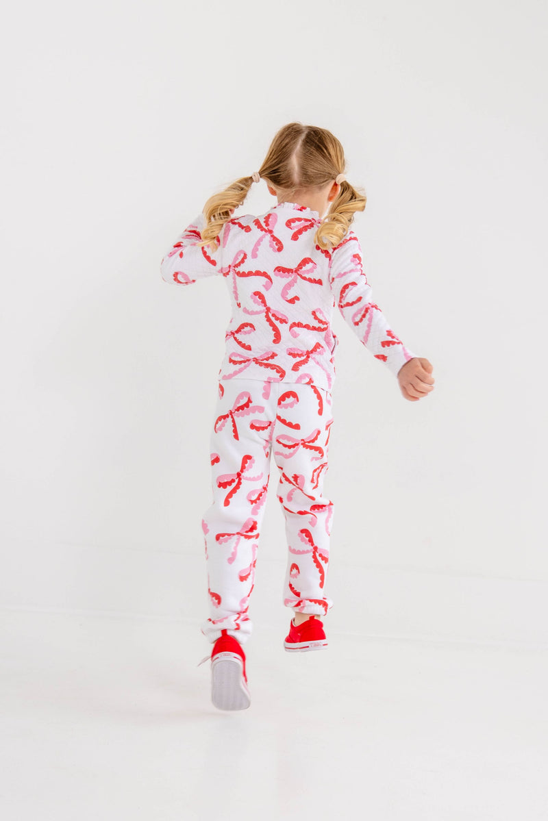 A child with pigtails seen from behind, wearing a long-sleeved shirt and Gates Sweeney Sweatpants - Bonnie Scot Bows with red and pink bows, paired with red sneakers.