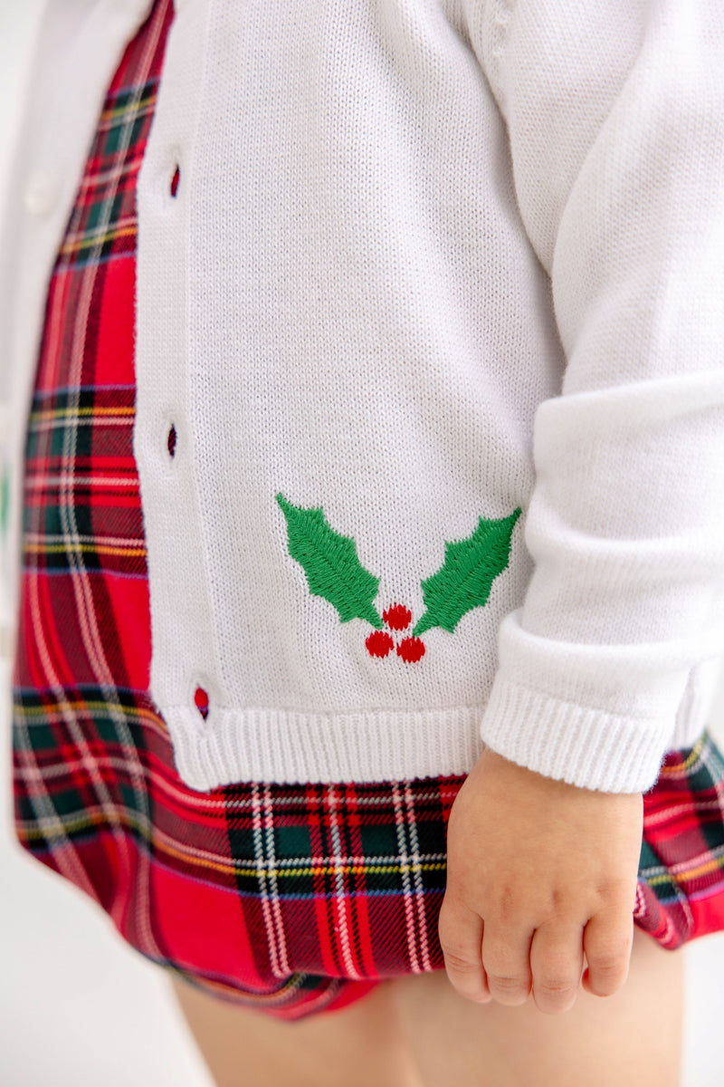 A child is shown wearing The Beaufort Bonnet Company’s Cambridge Cardigan - Worth Avenue White with Holly, featuring embroidered holly leaves and red berries, layered over a red plaid dress. Only the lower half of the child’s face and body is visible.