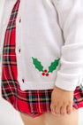 A child is shown wearing The Beaufort Bonnet Company’s Cambridge Cardigan - Worth Avenue White with Holly, featuring embroidered holly leaves and red berries, layered over a red plaid dress. Only the lower half of the child’s face and body is visible.